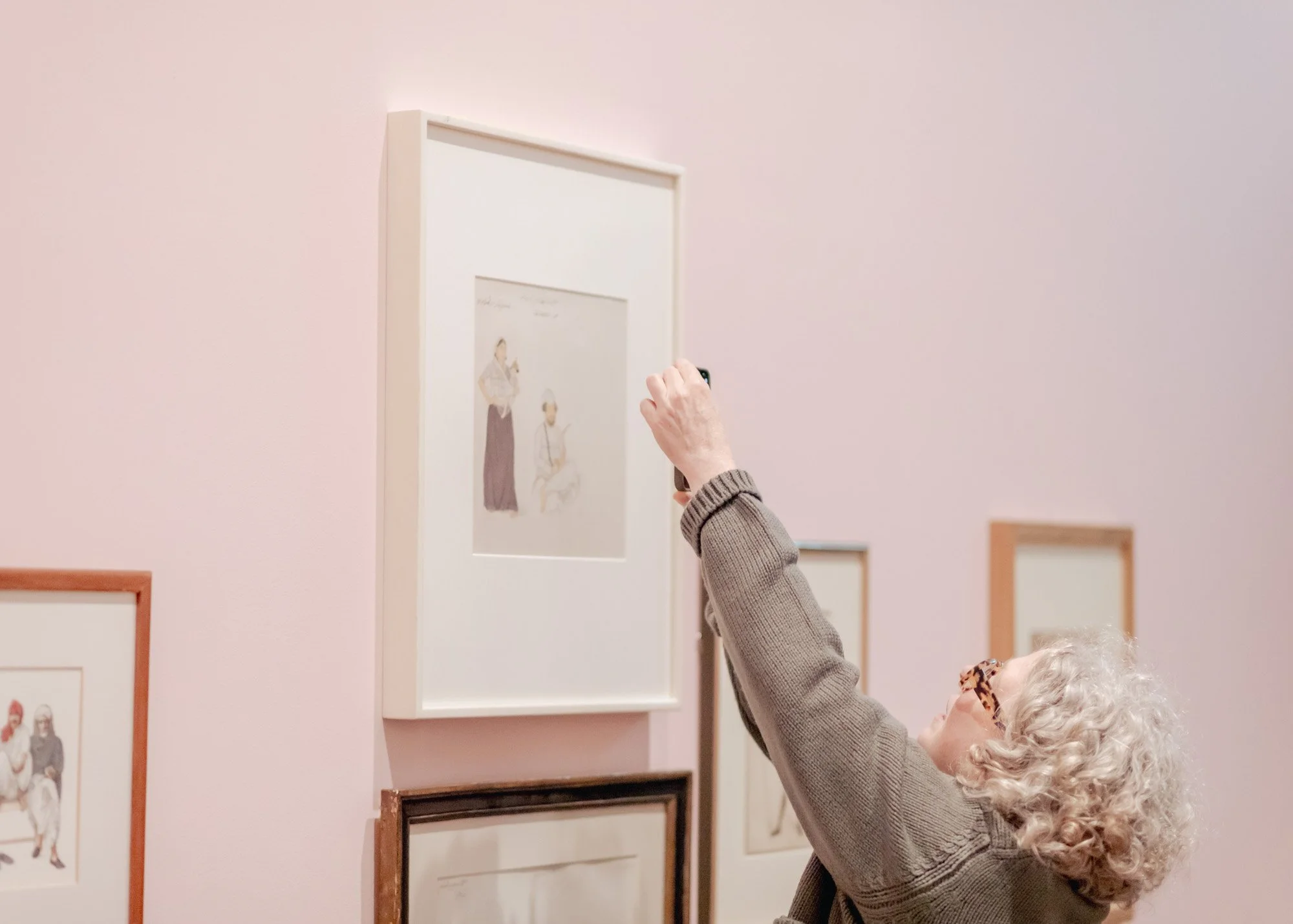 An elderly woman with white curly hair and leopard print glasses is taking a photo of a painting at an art gallery. She is wearing a gray cardigan.