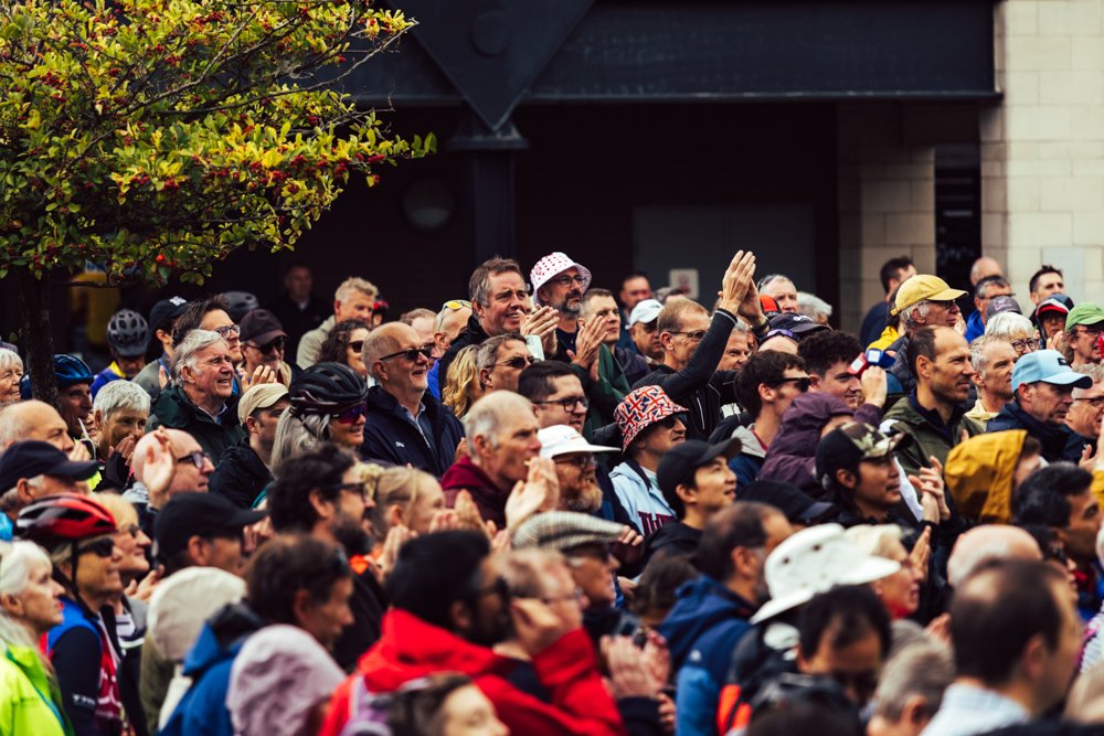 Crowd of people gathered outdoors, many wearing jackets, some clapping and smiling, with a tree and building in the background.