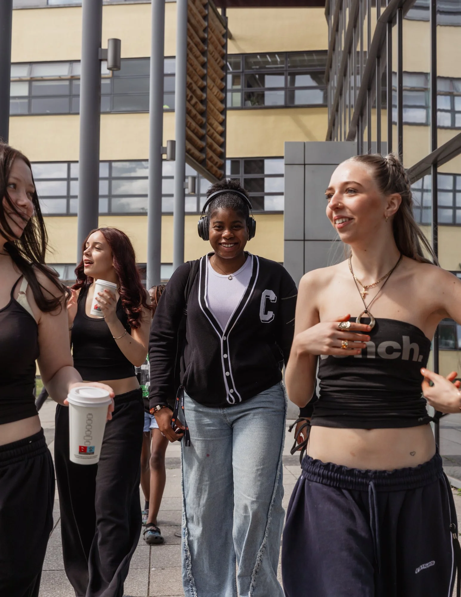 Group of young women walking and chatting outdoors near a modern building, with some holding coffee cups.