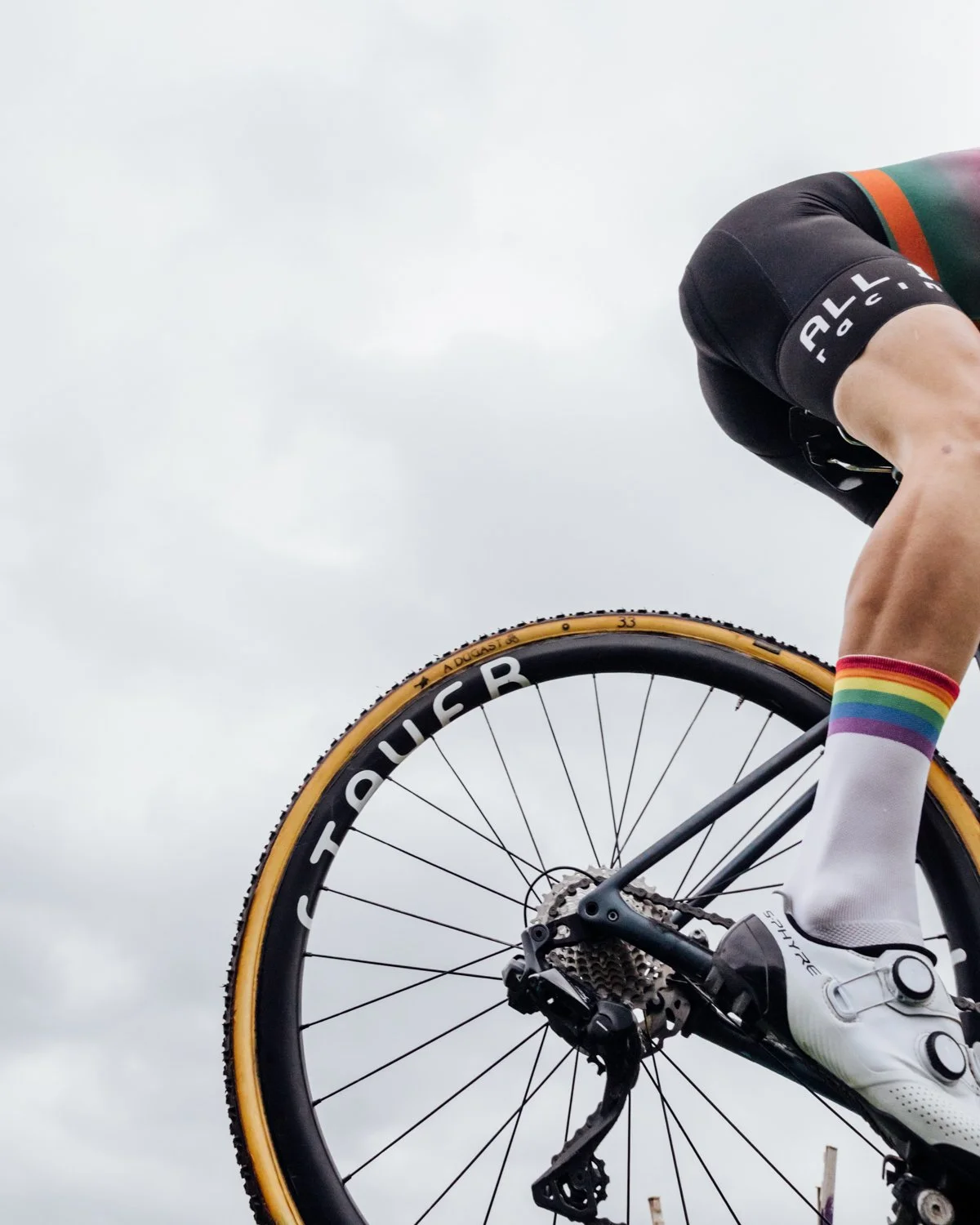 Close-up of a cyclist's lower body and bike, showing part of the rider's leg, colorful rainbow socks, white cycling shoes, and the rear wheel with a black tire labeled 'EC90'. The rider is wearing black shorts and is riding against a cloudy sky.
