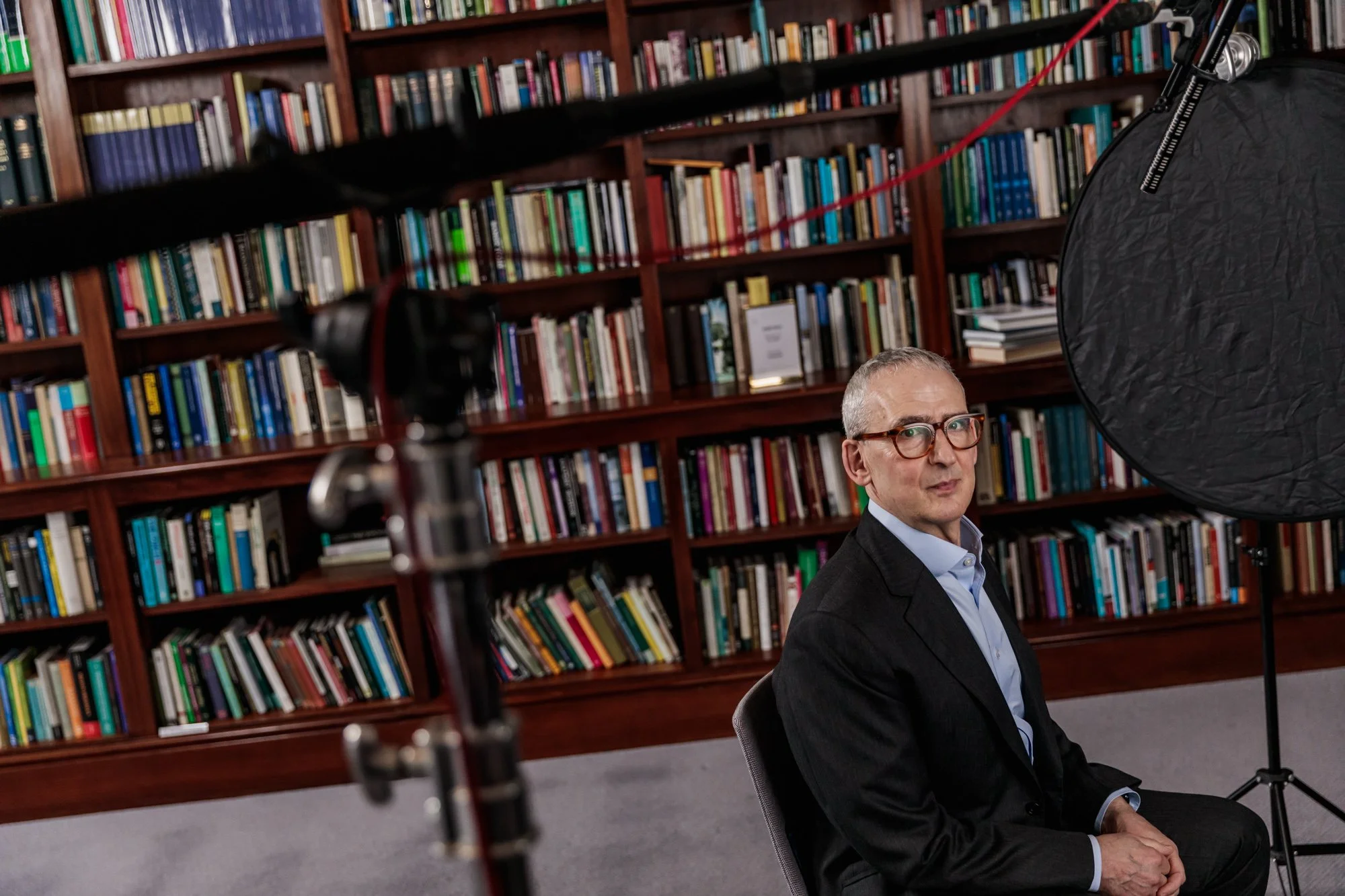 A man in a suit and glasses sitting in front of a bookshelf during a video recording session, with professional lighting equipment visible in the foreground.