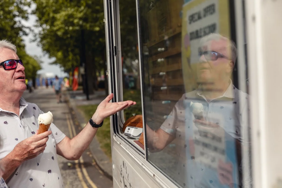 An older man with gray hair and glasses, wearing a light-colored polo shirt with small floral patterns, is holding an ice cream cone in his left hand and gesturing with his right hand towards a window of a mobile food truck. The reflection of the man