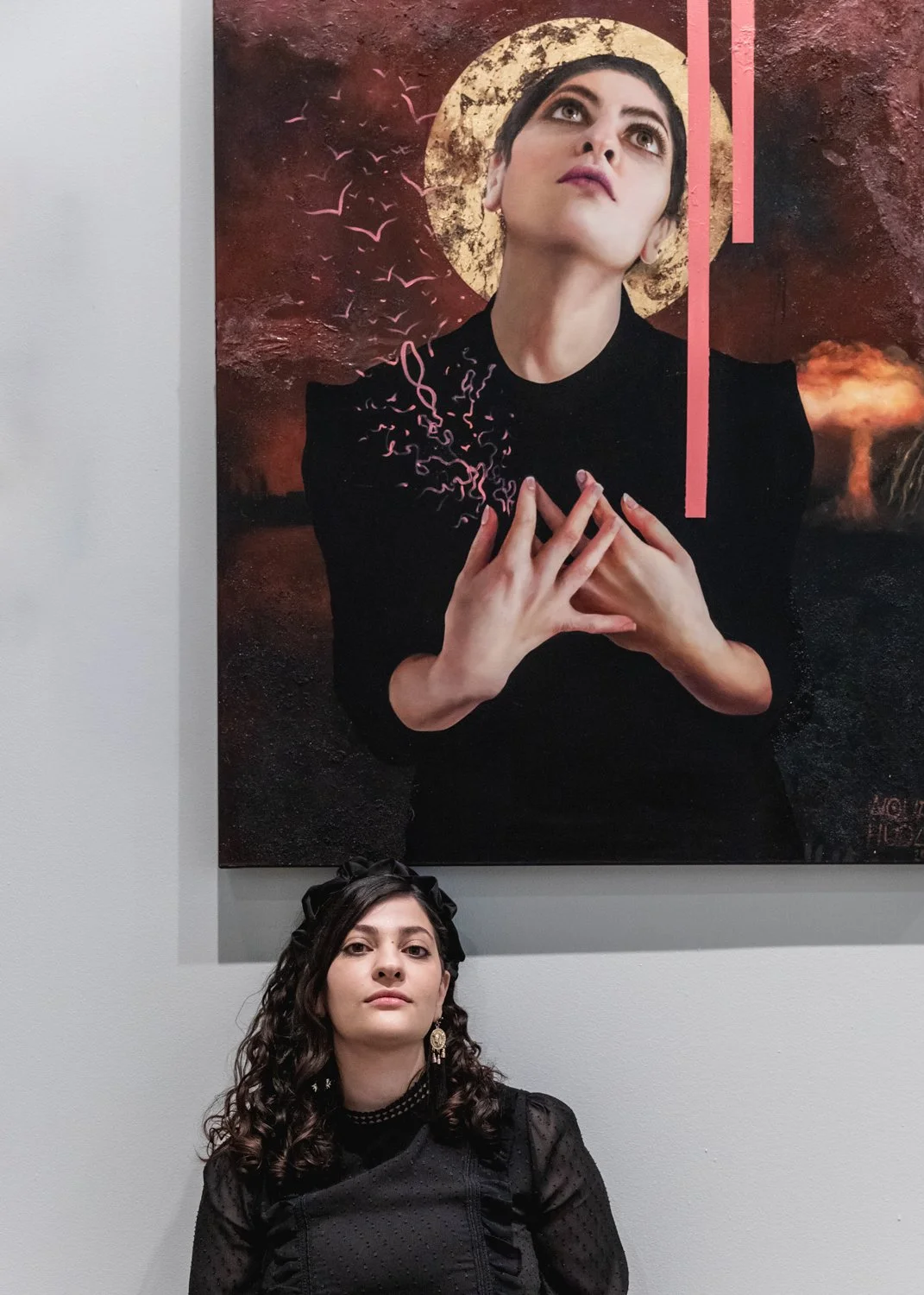 A woman with dark, curly hair, wearing black clothing and earrings, is sitting in front of a large painting. The painting depicts a woman with short hair, with her hands placed on her chest, against a dark, textured background with a large moon and p