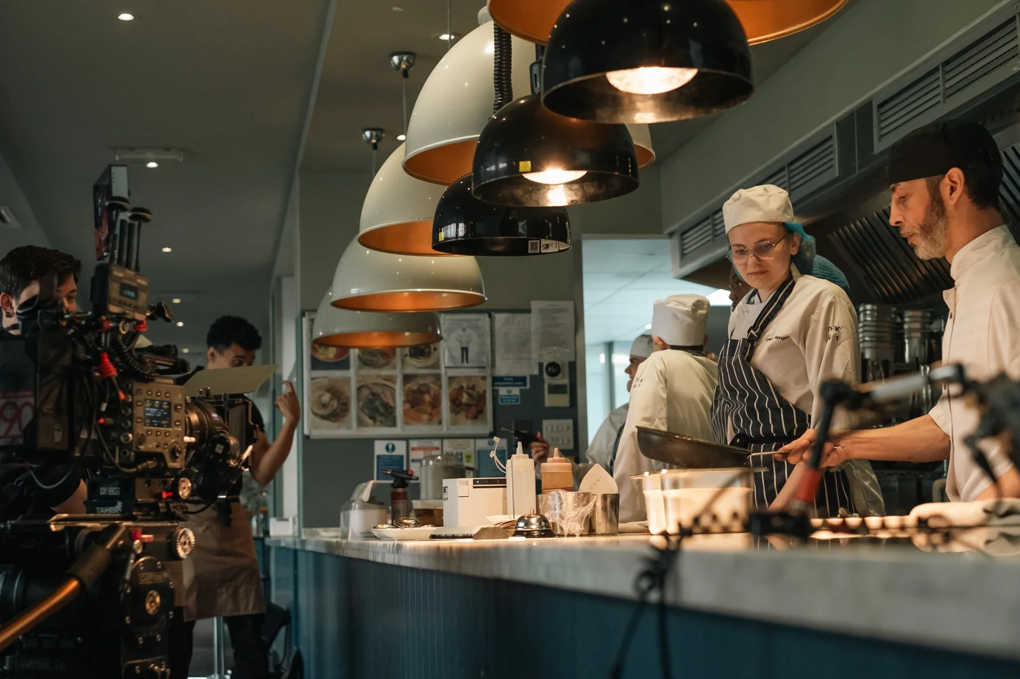 A professional kitchen where chefs and a film crew are filming a cooking show. Chefs are preparing dishes, with one holding a pan, while crew members operate cameras.