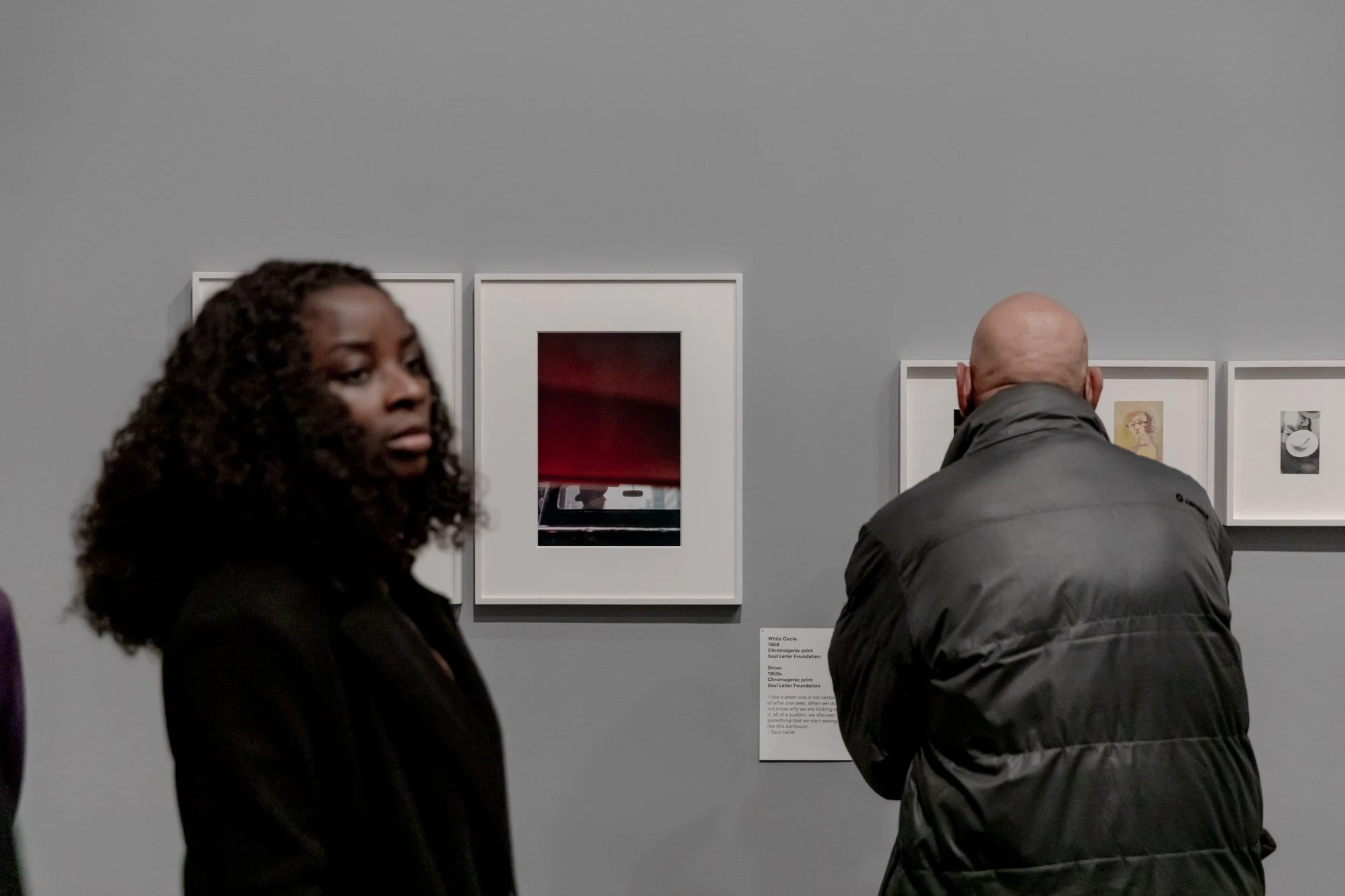 Two people viewing artwork in a gallery. The woman in the foreground has curly dark hair and is looking to the side, while the man in the background, with a bald head, is wearing a black jacket and is looking at the artwork on the wall.