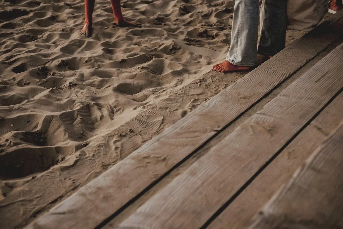 Close-up of sandy beach with footprints and a person in jeans and a white shirt standing near a wooden walkway.