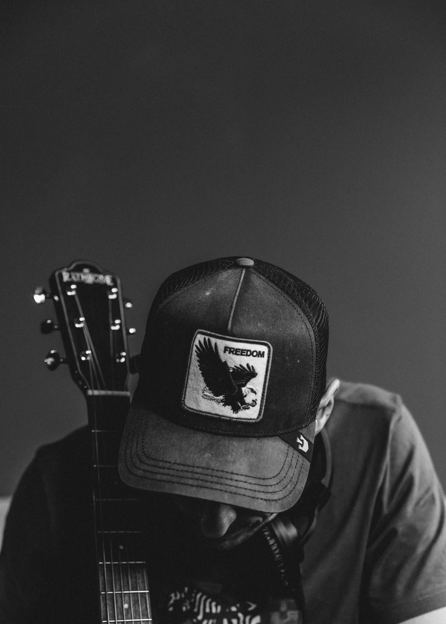 A person wearing a cap with an eagle patch labeled 'FREEDOM', obscuring part of their face, holding an acoustic guitar against a plain background.