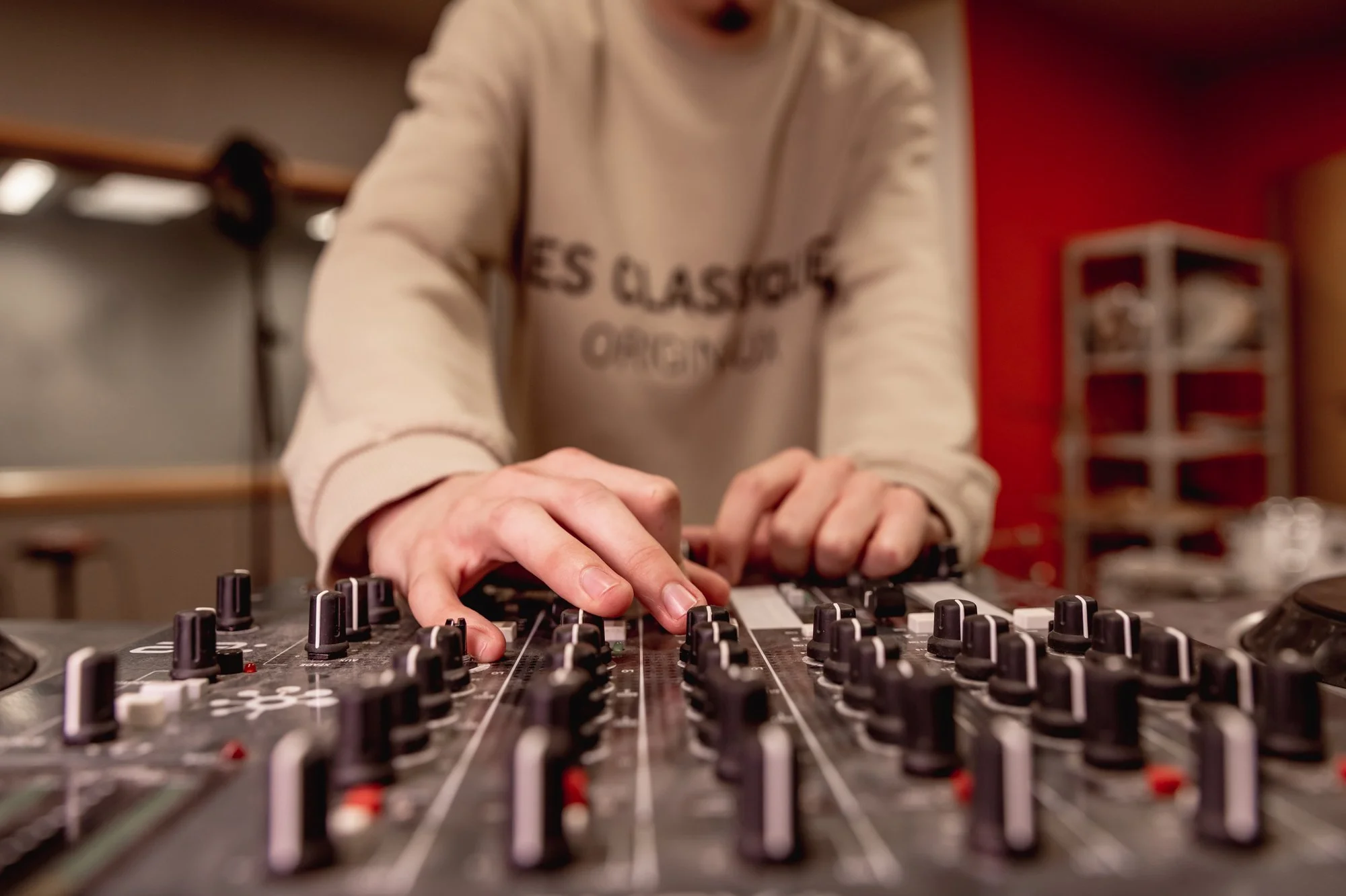 Person adjusting controls on a DJ mixing console in a room with red walls and shelves in the background.