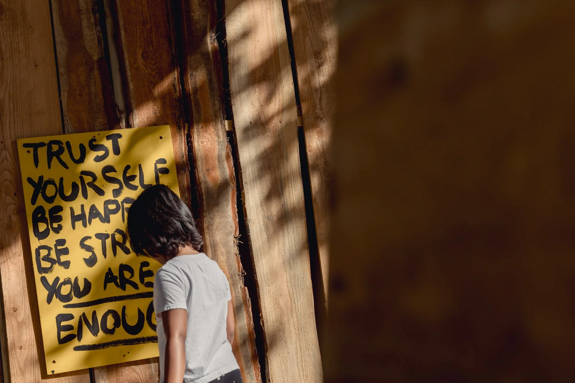 A young child with dark hair wearing a white T-shirt is standing in front of a wooden wall, looking at a large yellow poster with black handwritten text that reads 'Trust Yourself Be Happy Be Strong You Are Enough.'