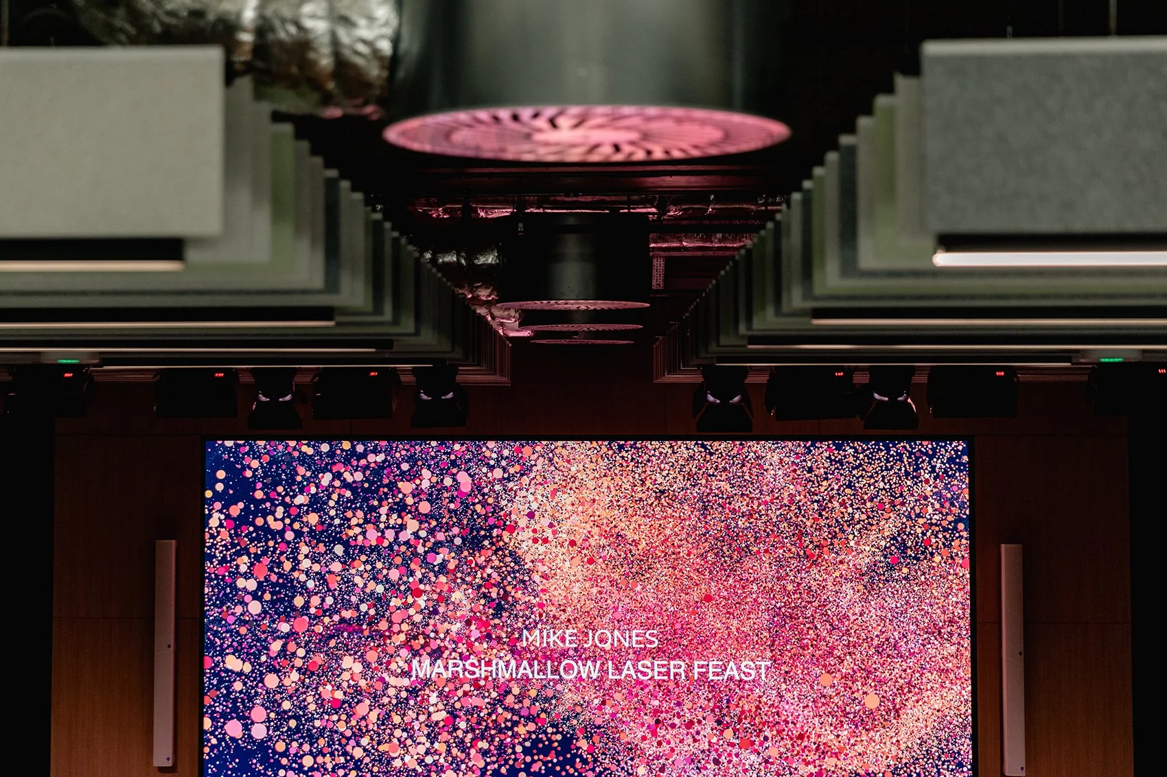 A large digital screen displays the text 'MIKE JONES MARSHMALLOW LASER FEAST' against a vibrant, colorful background of pink, purple, and white dots resembling a cosmic or confetti pattern. Above the screen, stage lighting fixtures and black ceiling 