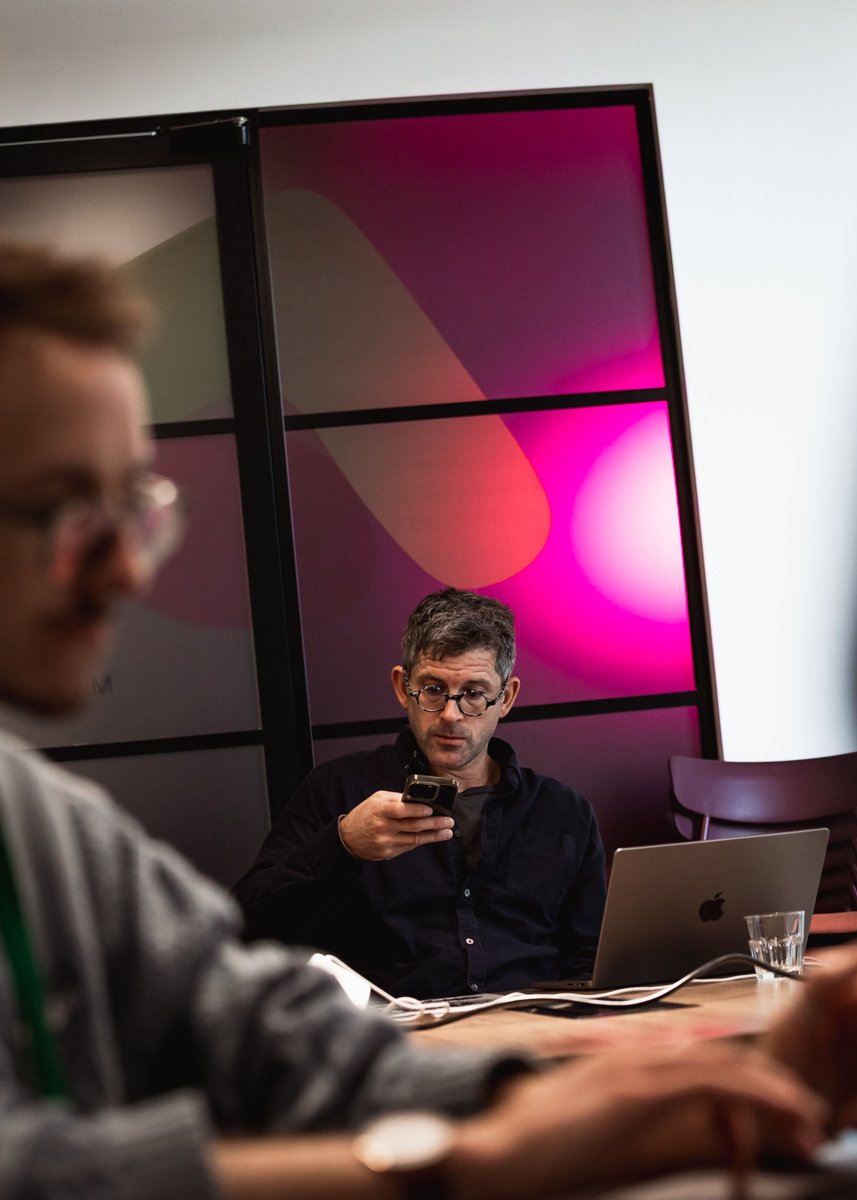 Man with glasses sitting at a table with a laptop, holding a smartphone, in a meeting room with a colorful abstract background.