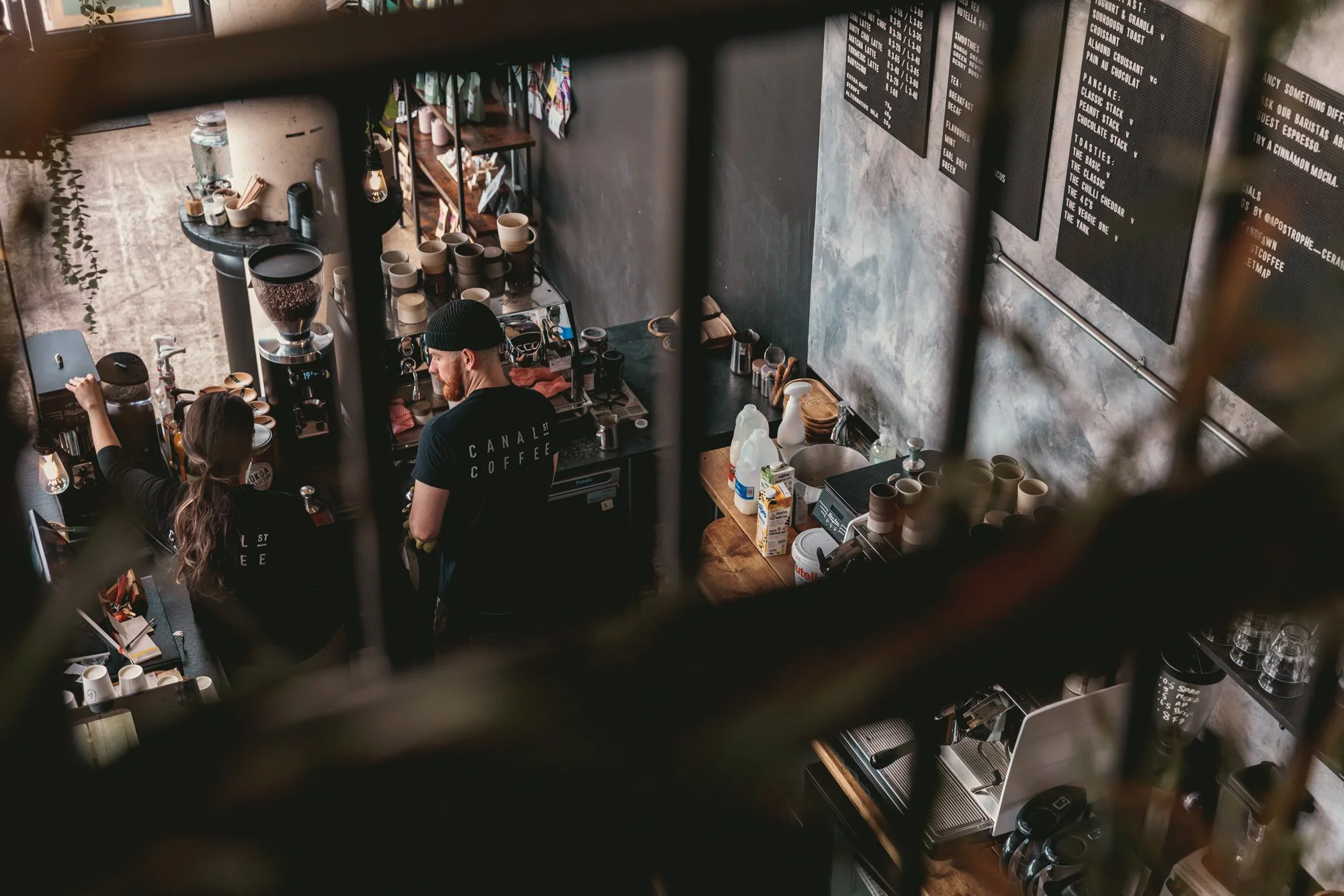 Two baristas behind the counter in a coffee shop, one male and one female, preparing coffee. The shop has a modern industrial style with a black menu board on the wall and various coffee equipment and supplies on the counters.