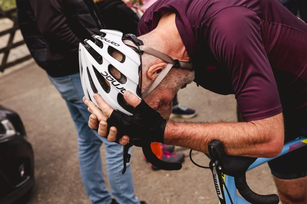A man in a maroon cycling jersey is bent over, holding his head with his hands, wearing a white bicycle helmet with black accents, while sitting on a blue bicycle.