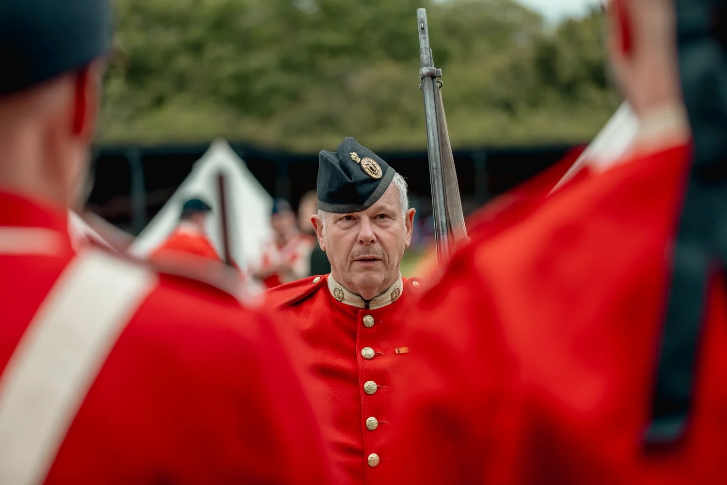 A man dressed in a historical military uniform, including a red coat with gold buttons and a black hat with an emblem, stands among other men in similar attire during a reenactment or historical event. The background features tents and trees.
