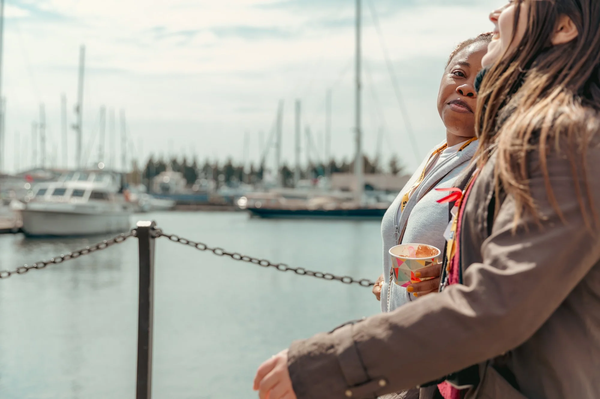 Two women standing near a marina with boats, one holding a colorful cup, talking and smiling.
