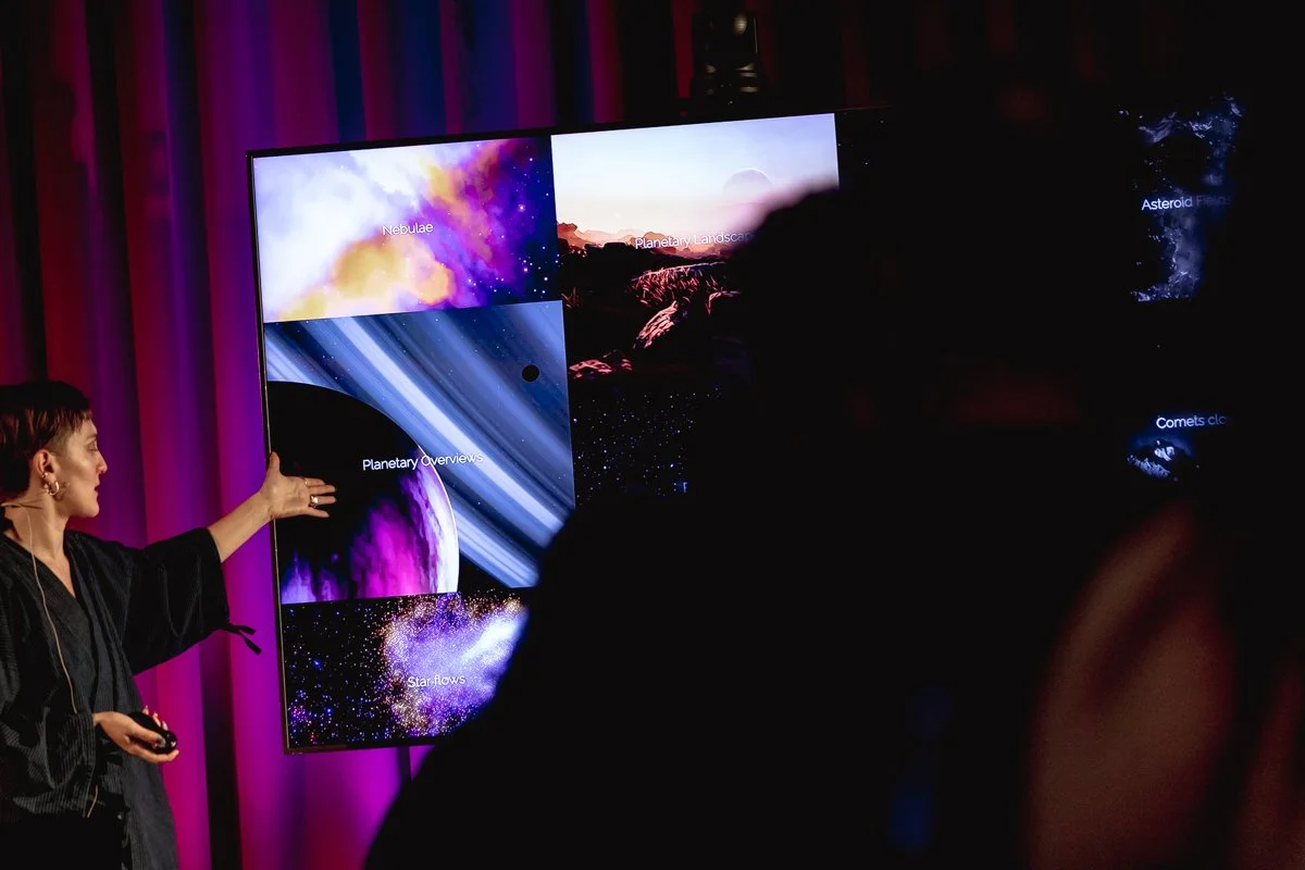 A woman presenting information about space on a large display screen, with spotlights and a dark background.