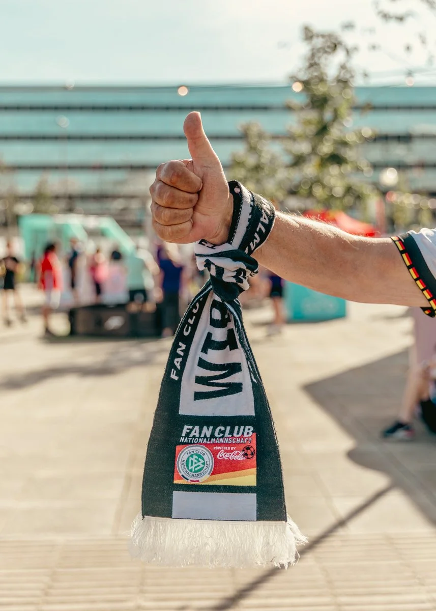 A person holding a black and white scarf with the words 'FAN CLUB' and the logo of a football team, in an outdoor setting with people and food trucks in the background.