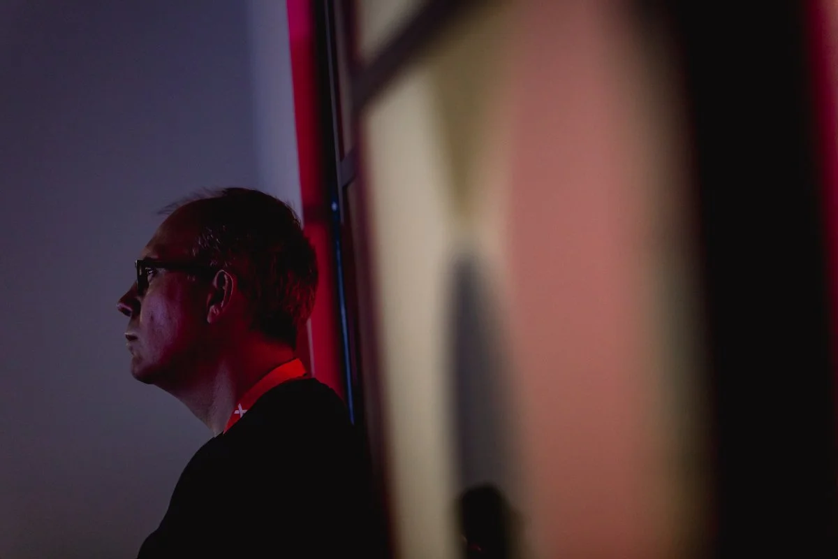 A man wearing glasses and a red lanyard looking to the left against a dark background, illuminated by red and purple lighting, partially obscured by an out-of-focus object in the foreground.