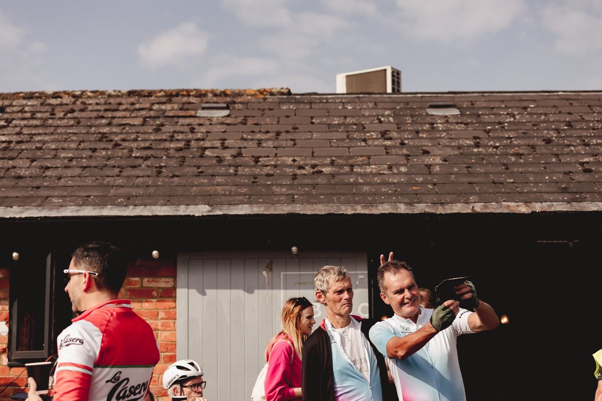 A group of people, most wearing cycling gear, are gathered outside a building with a gray door. One man takes a selfie, and others are standing around, including a woman in a pink jacket and some men in cycling jerseys and helmets.