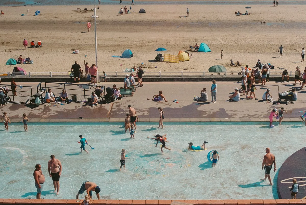 People enjoying a day at a beach with a swimming pool in the foreground, sandy beach with umbrellas, tents, and beachgoers in the background.