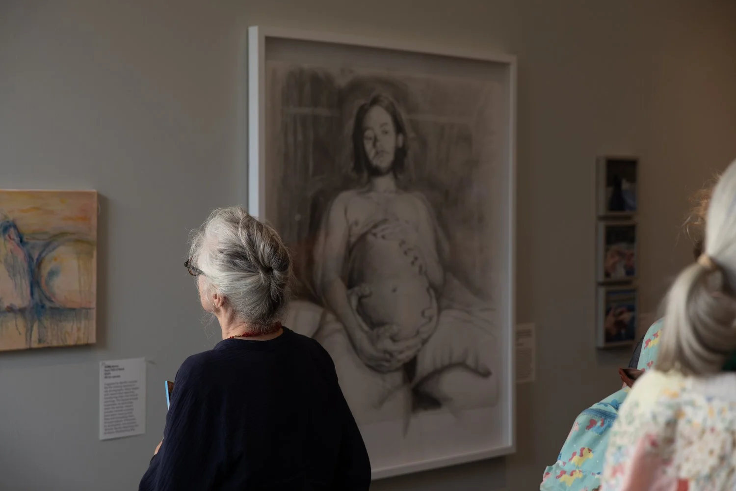 Visitors at an art gallery viewing a large black-and-white sketch of a woman with short hair, sitting with her hands on her knee, holding her belly, with other paintings displayed on the wall.