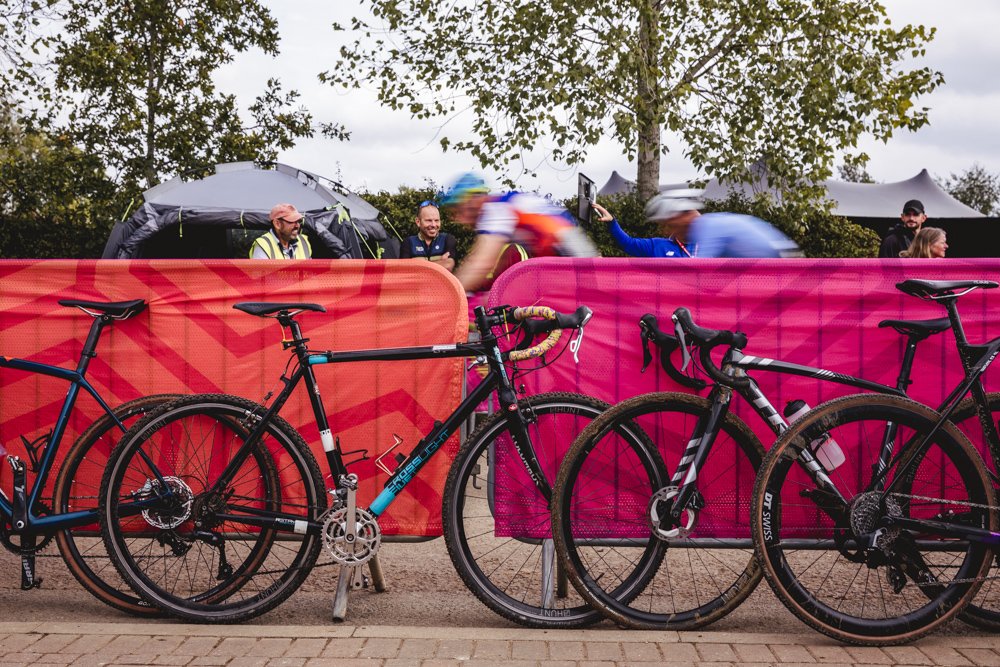 Bicycles parked against pink barriers at a cycling event, with blurred cyclists and spectators in the background under cloudy sky.