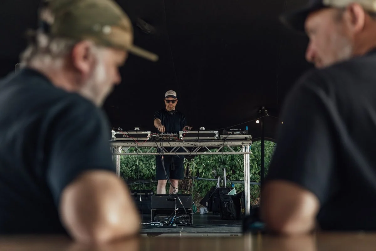 DJ performing on stage at an outdoor event, with two men in black shirts and hats in the foreground.