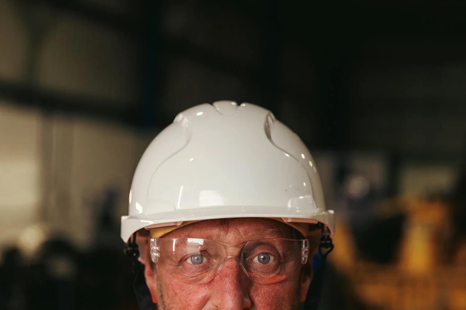 Close-up of a man wearing a white safety helmet and clear safety glasses in industrial setting.