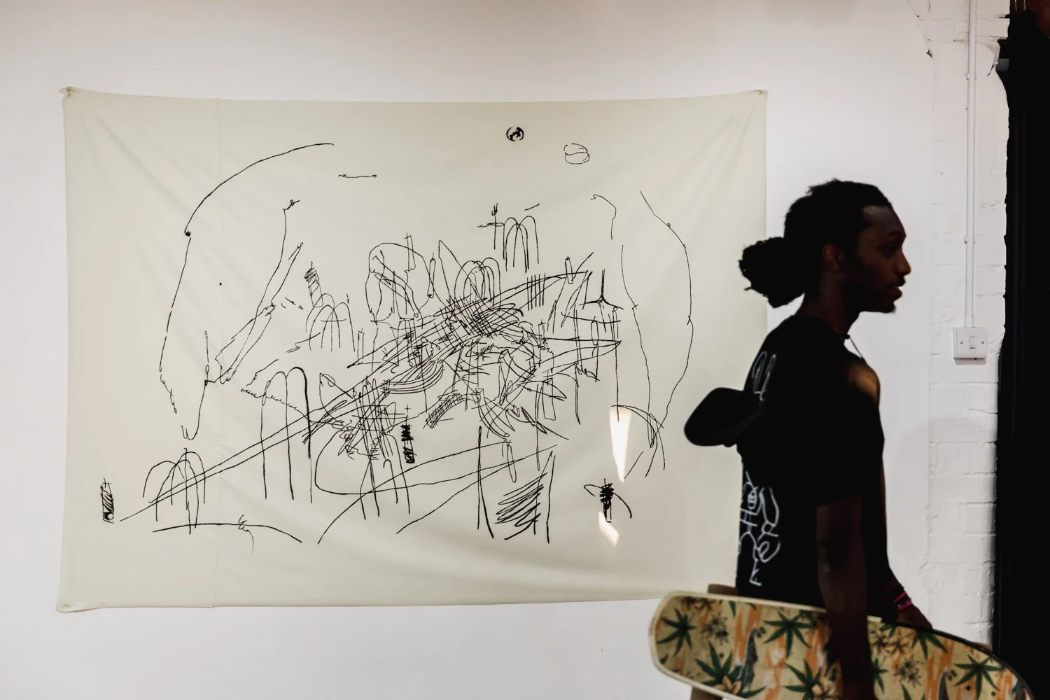 A man holding a skateboard with cannabis leaf patterns in front of an abstract black line drawing on a white wall.