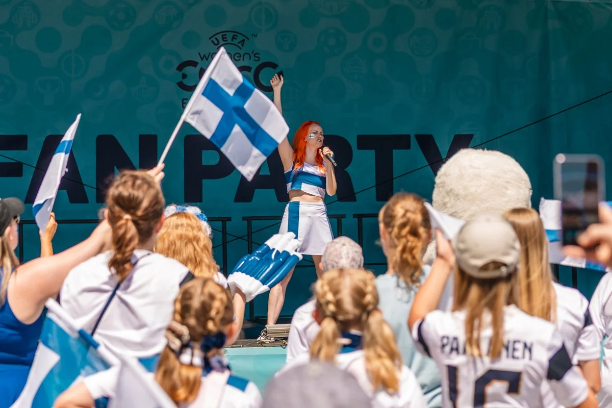 A woman with red hair and face paint is speaking into a microphone on stage during a women's sports event, surrounded by children and spectators holding Finnish flags, with a large sign that reads 'UEFA Women's Euro' in the background.