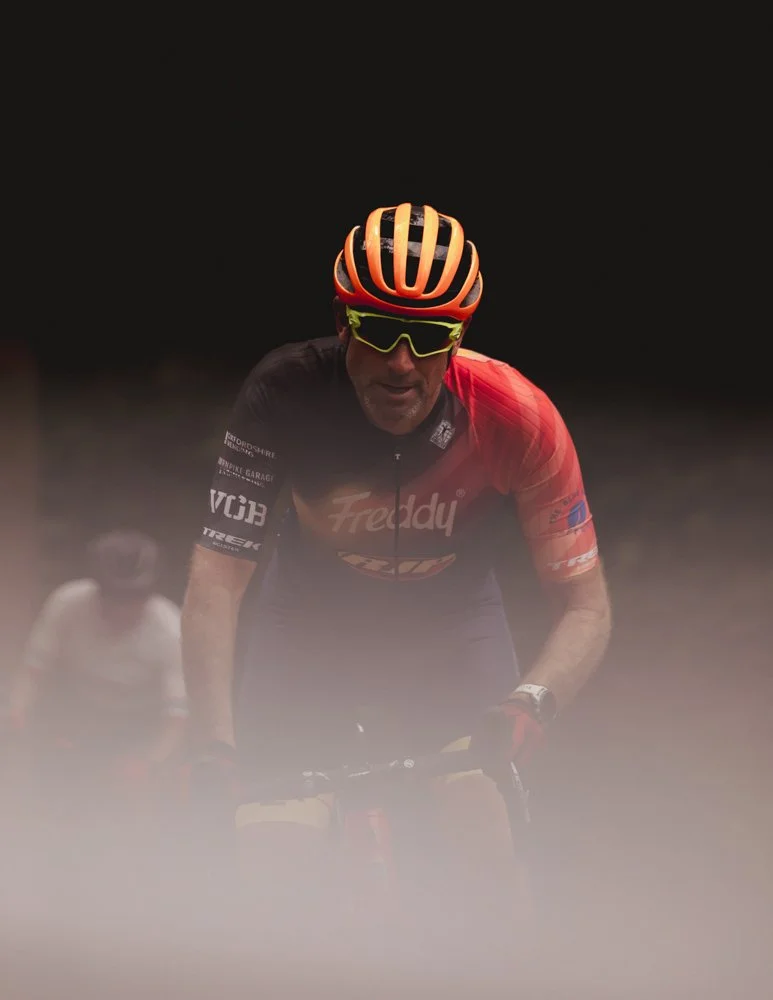A cyclist wearing an orange helmet, yellow sunglasses, and a black and red jersey riding a mountain bike during a race at night, with another cyclist visible in the background.
