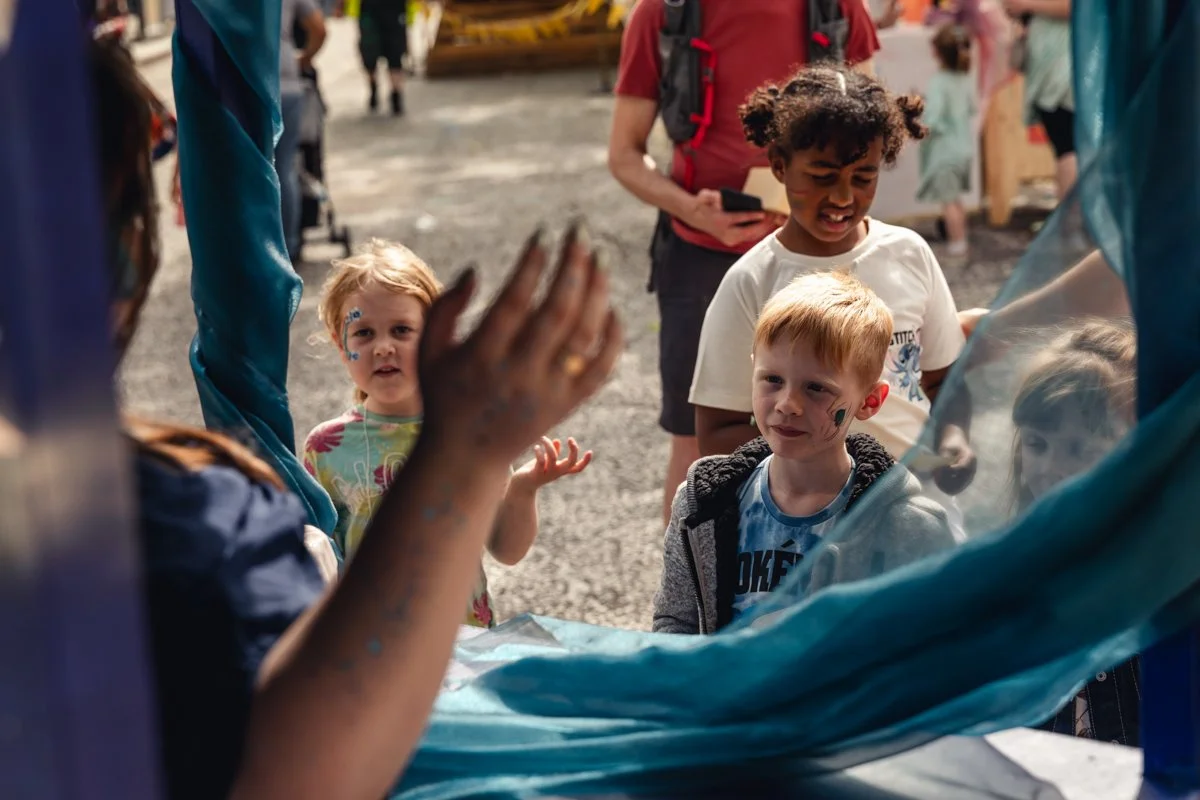 A group of children talking to an adult through a blue plastic barrier at an outdoor event, with other tents and people in the background.