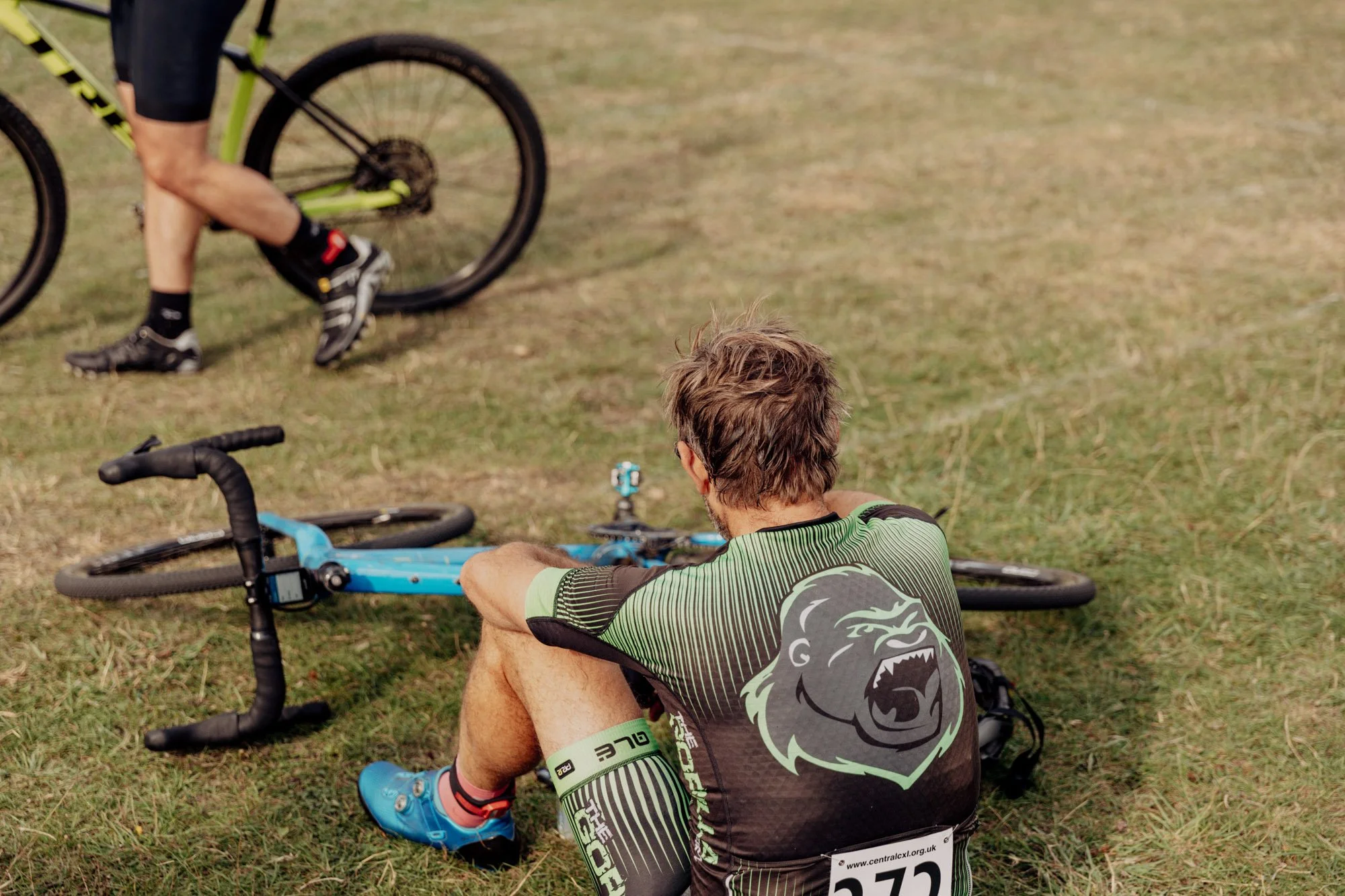 A man sitting on the grass next to a fallen blue bicycle, resting with his arms on his knees, after a cycling event.