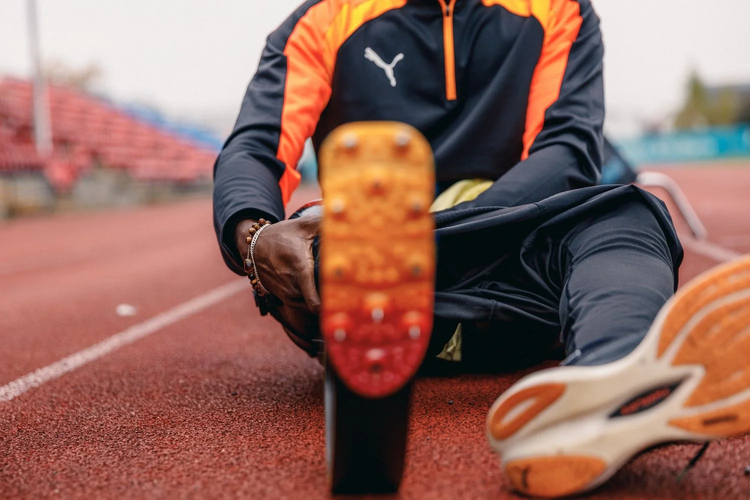 Athlete in black and orange Puma sportswear sitting on track, focusing on shoe with blurred background of stadium seating.