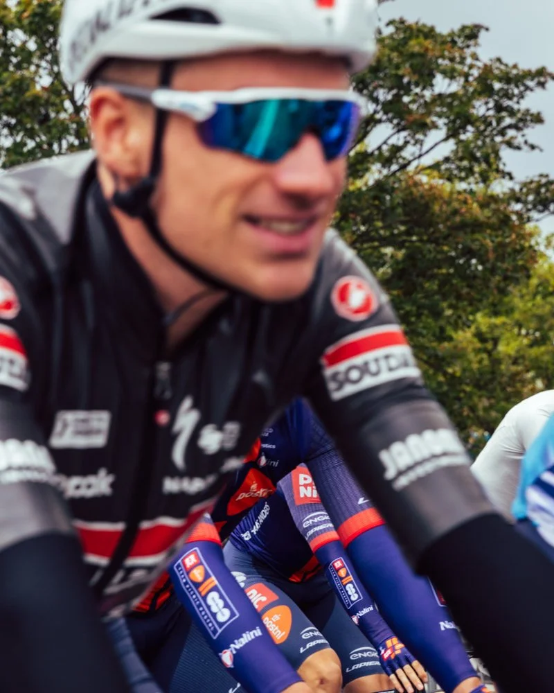Close-up of a cyclist wearing a white helmet with black straps, reflective blue sunglasses, and a black and red cycling jersey, during a race or training outdoors with trees in the background.