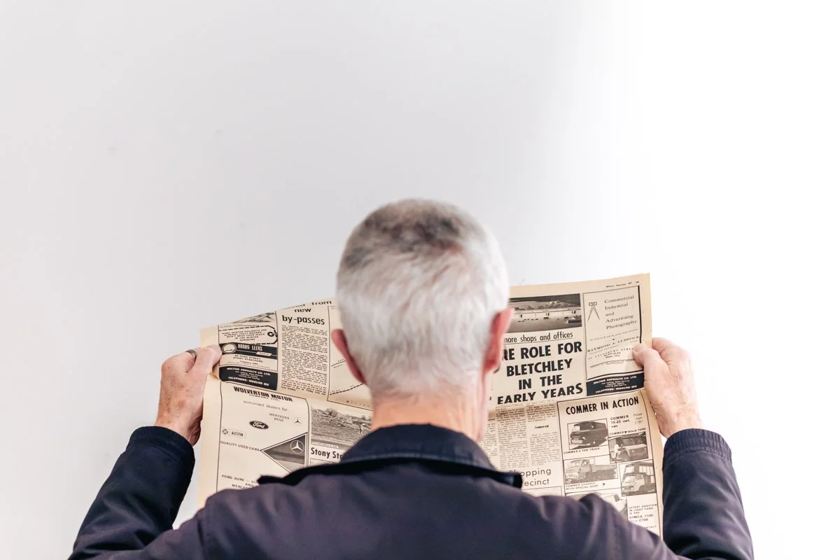 A man with gray hair wears a dark jacket and reads a newspaper against a plain white wall.