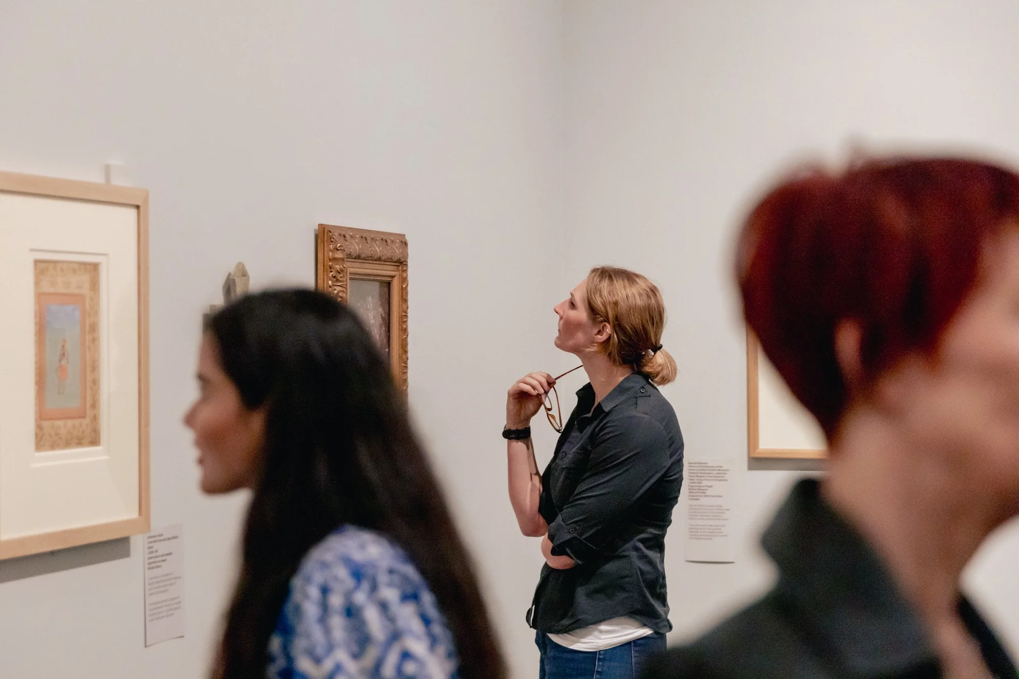 People viewing art in a museum, focusing on a woman with reddish hair, black shirt, and glasses, standing with hand on chin, surrounded by framed artwork on the white walls.