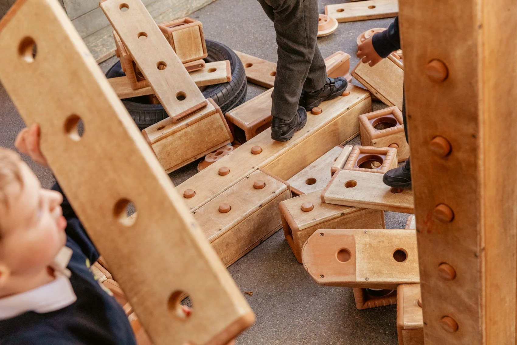 Children playing on a wooden block construction set, with large wooden pieces and rubber tires, on a gray carpeted surface.
