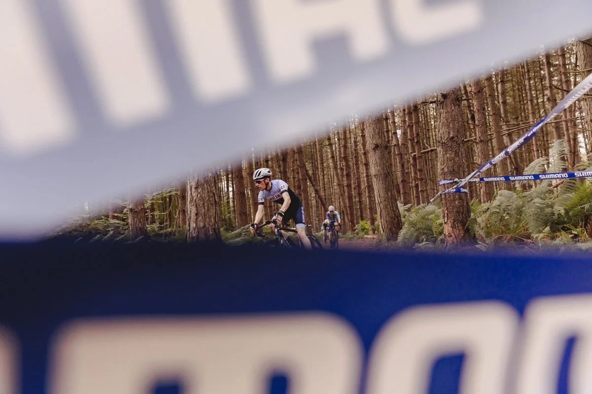 Cyclists riding mountain bikes through a forested trail, viewed through a blue and white barrier with the Shimano logo.
