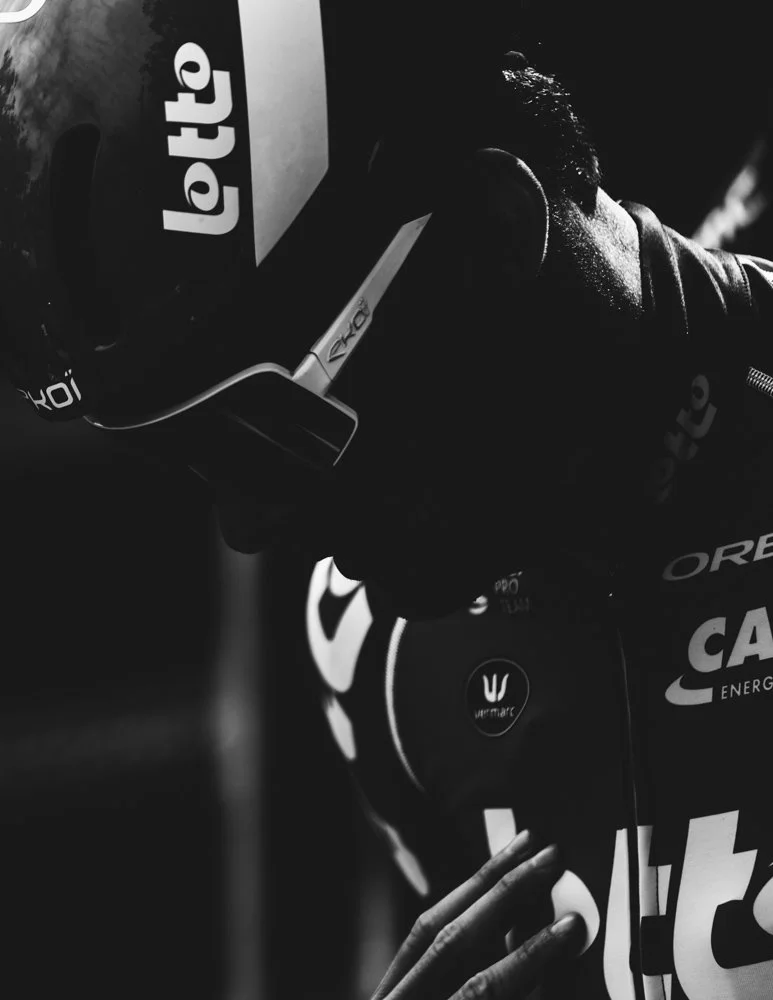 A close-up black and white photo of a person in a racing suit and helmet, leaning on a surface with various sponsor logos, including Nike, CA, and Umpart.