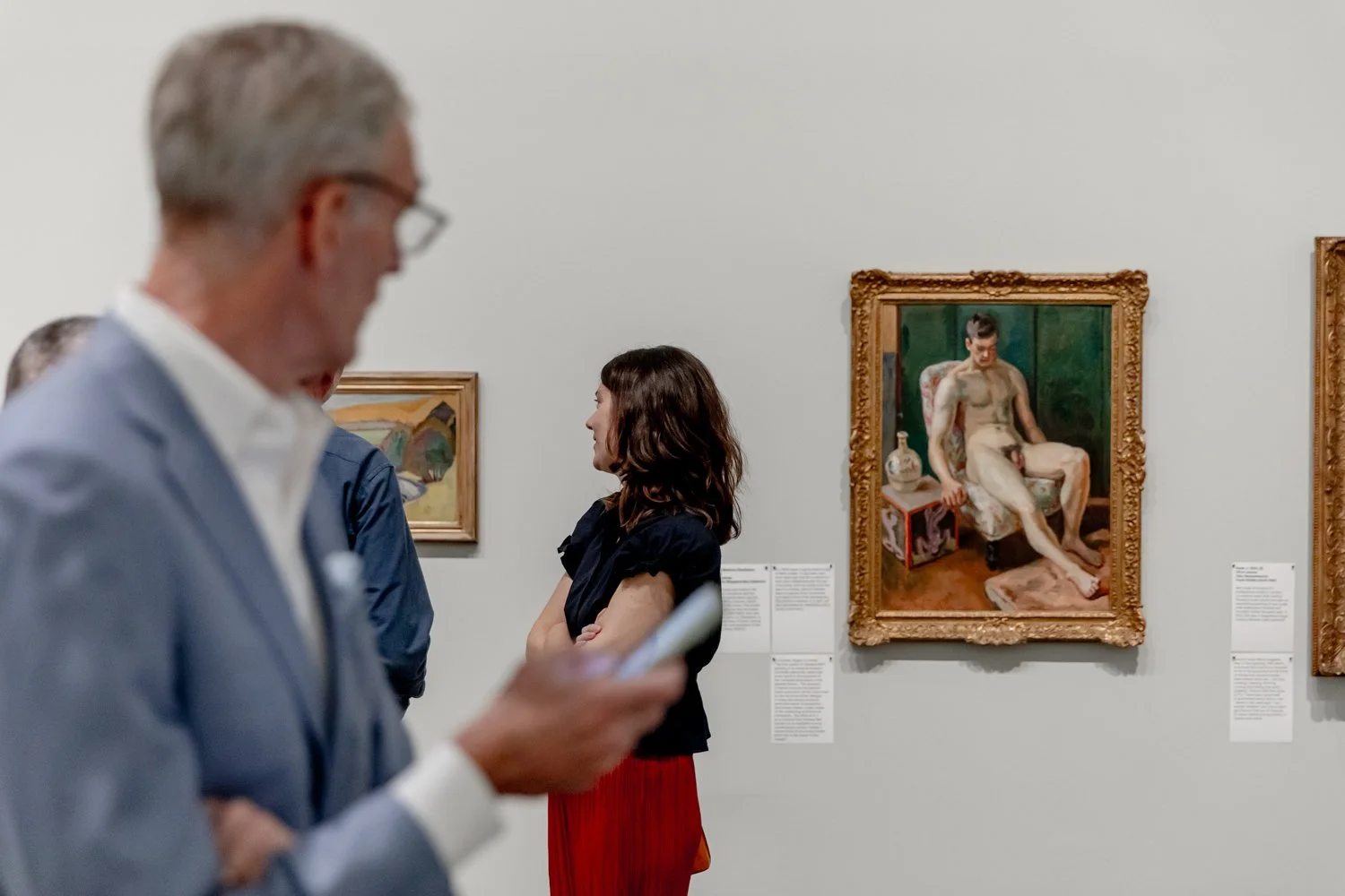 People viewing artwork in a gallery, with one man in the foreground looking at his phone and a woman in the background looking at paintings.