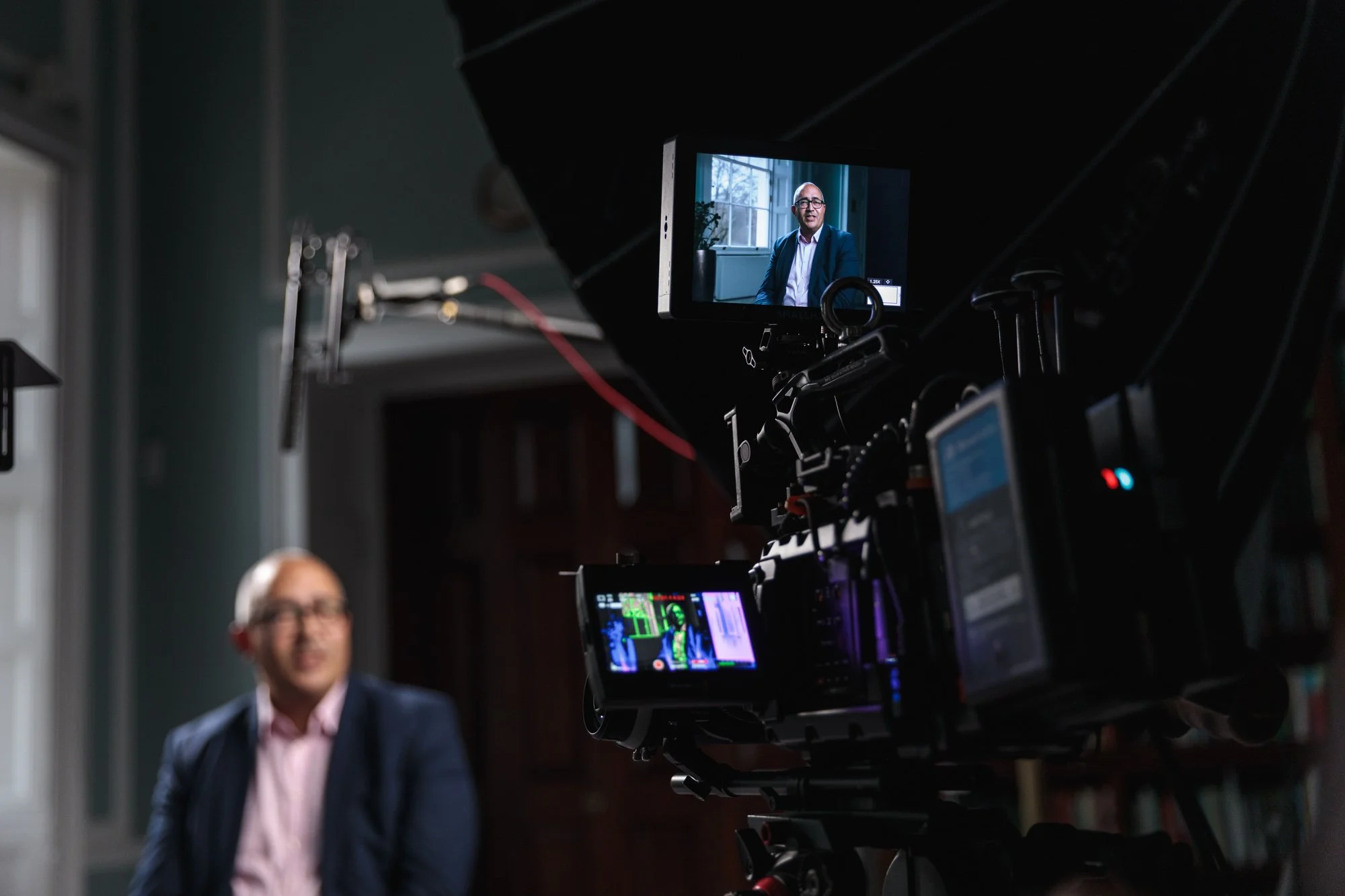 A man in a suit is being recorded for a video interview or presentation in a room with large windows and wood-paneled walls, seen through a professional video camera.