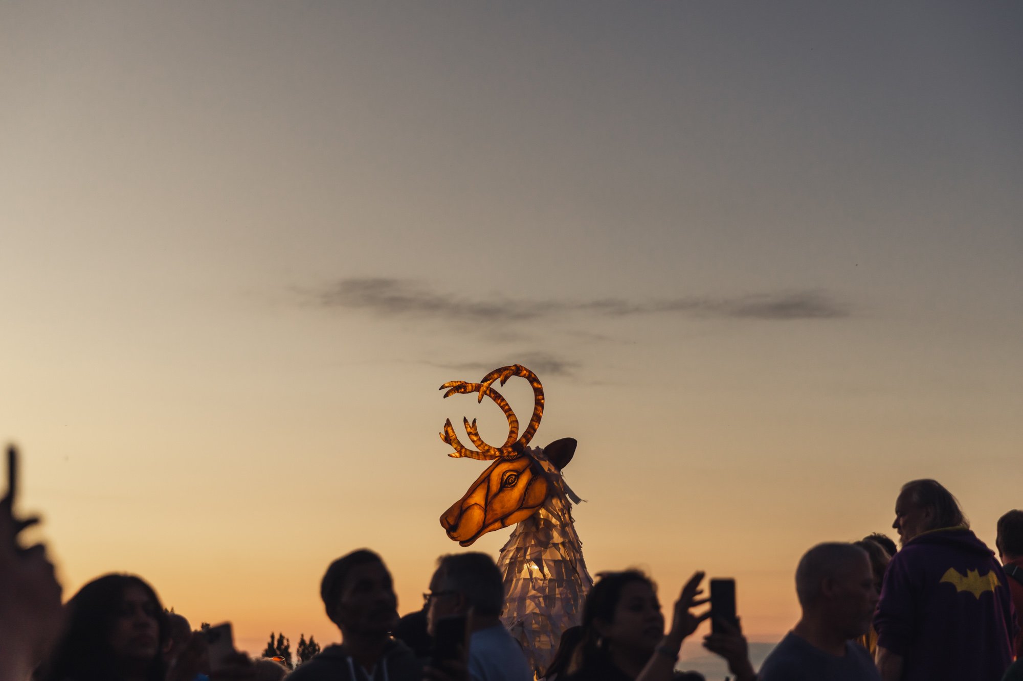 A large, illuminated reindeer puppet with antlers stands among a crowd of people at sunset, with some taking photos.