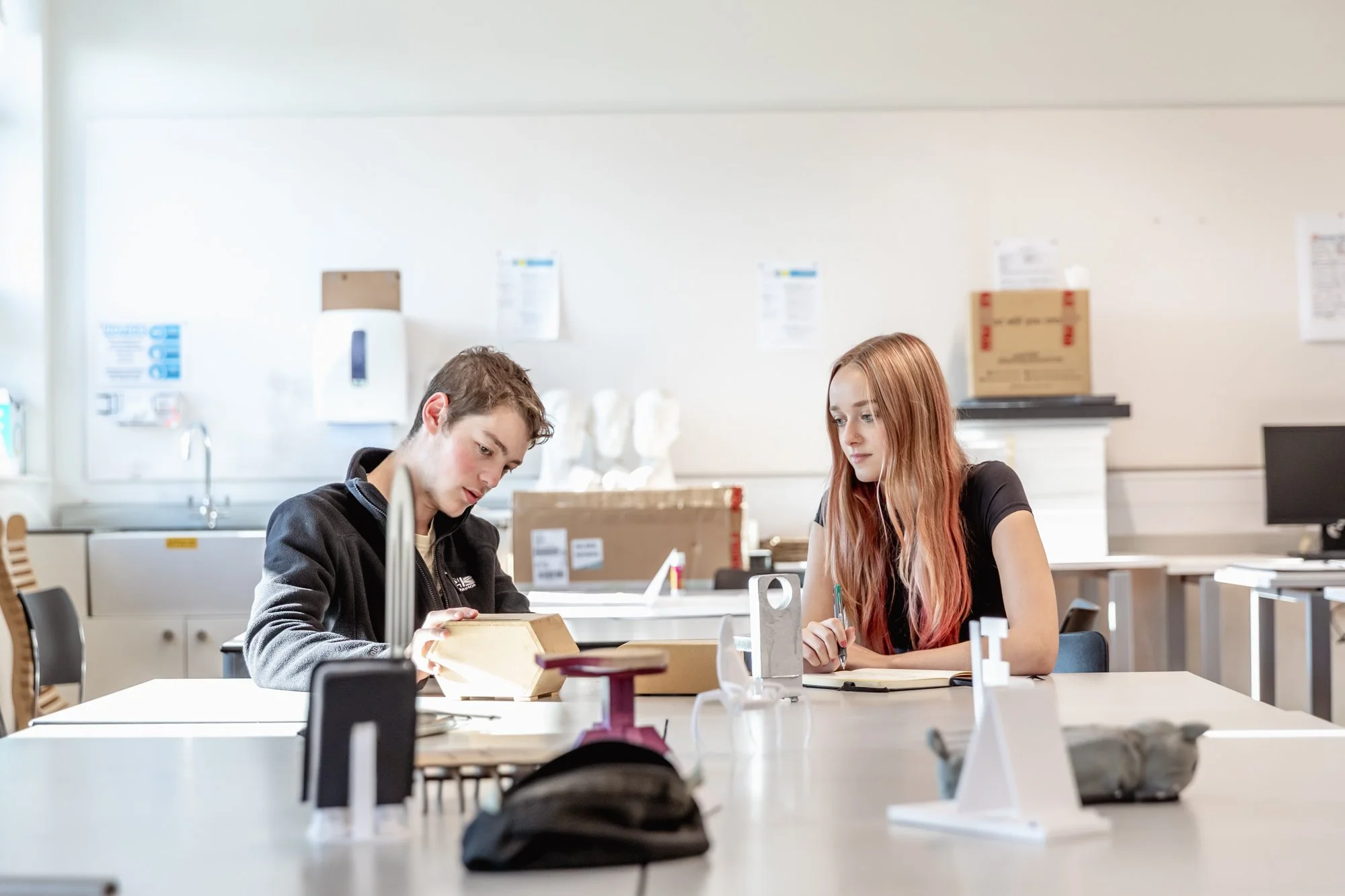 Two students, a young man and a young woman, sitting at a table in a classroom, studying and taking notes.