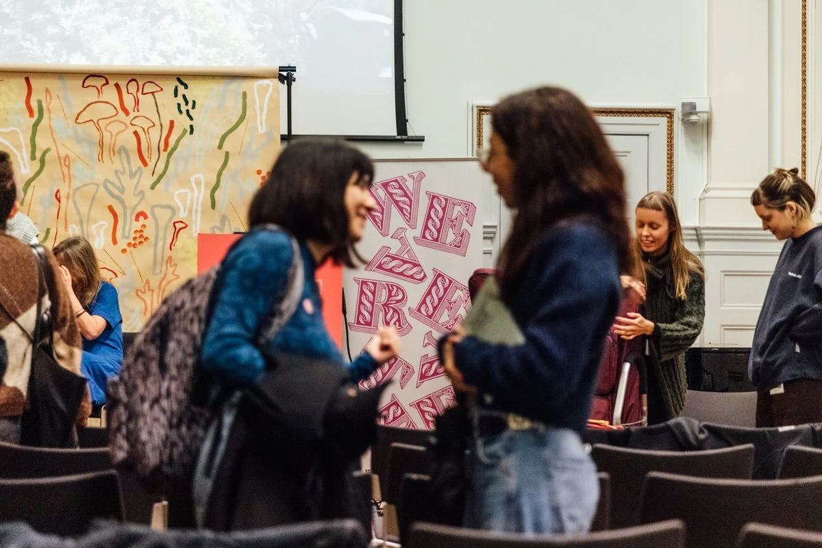 Two women are talking in a room with chairs, with one woman smiling and the other holding a phone. In the background, there are other people and colorful wall art with abstract patterns and the words "NSE".