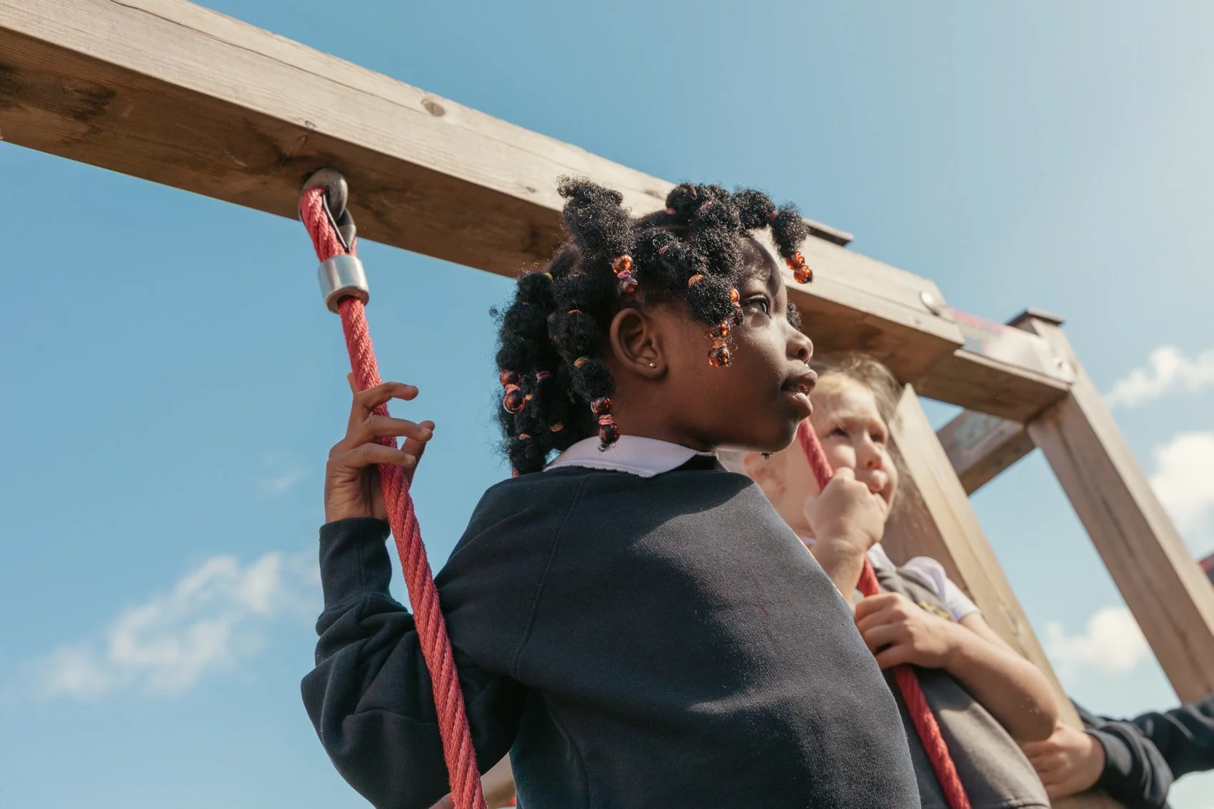 Two children, one girl with braided hair and beads, holding onto a red rope swing, standing against a blue sky with clouds.