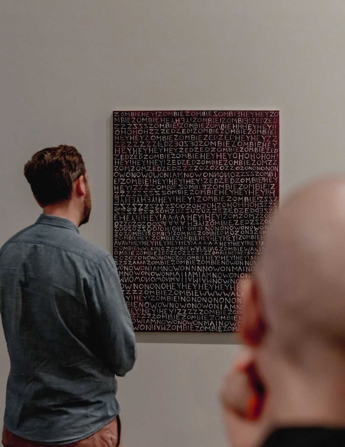 A man with short brown hair and a beard is standing in front of a wall showing a rectangular black and red artwork filled with white text and symbols. The man is wearing a gray shirt. Another person with blonde hair and earrings is partially visible 