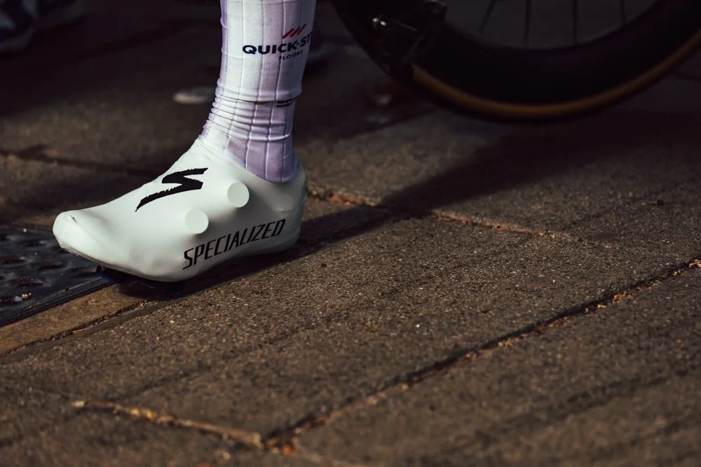 Close-up of a cyclist's foot wearing a white Specialized cycling shoe and long white socks, next to a bicycle wheel on a paved surface.