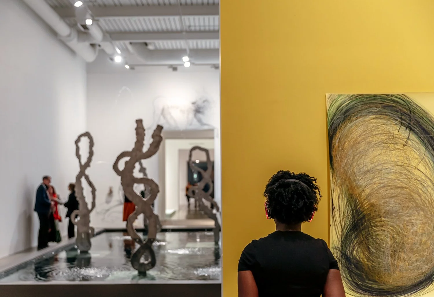 A woman with curly hair and headphones viewing abstract art in an art gallery. In the background, a white wall has sculptures and other artworks, with some visitors observing the exhibits.