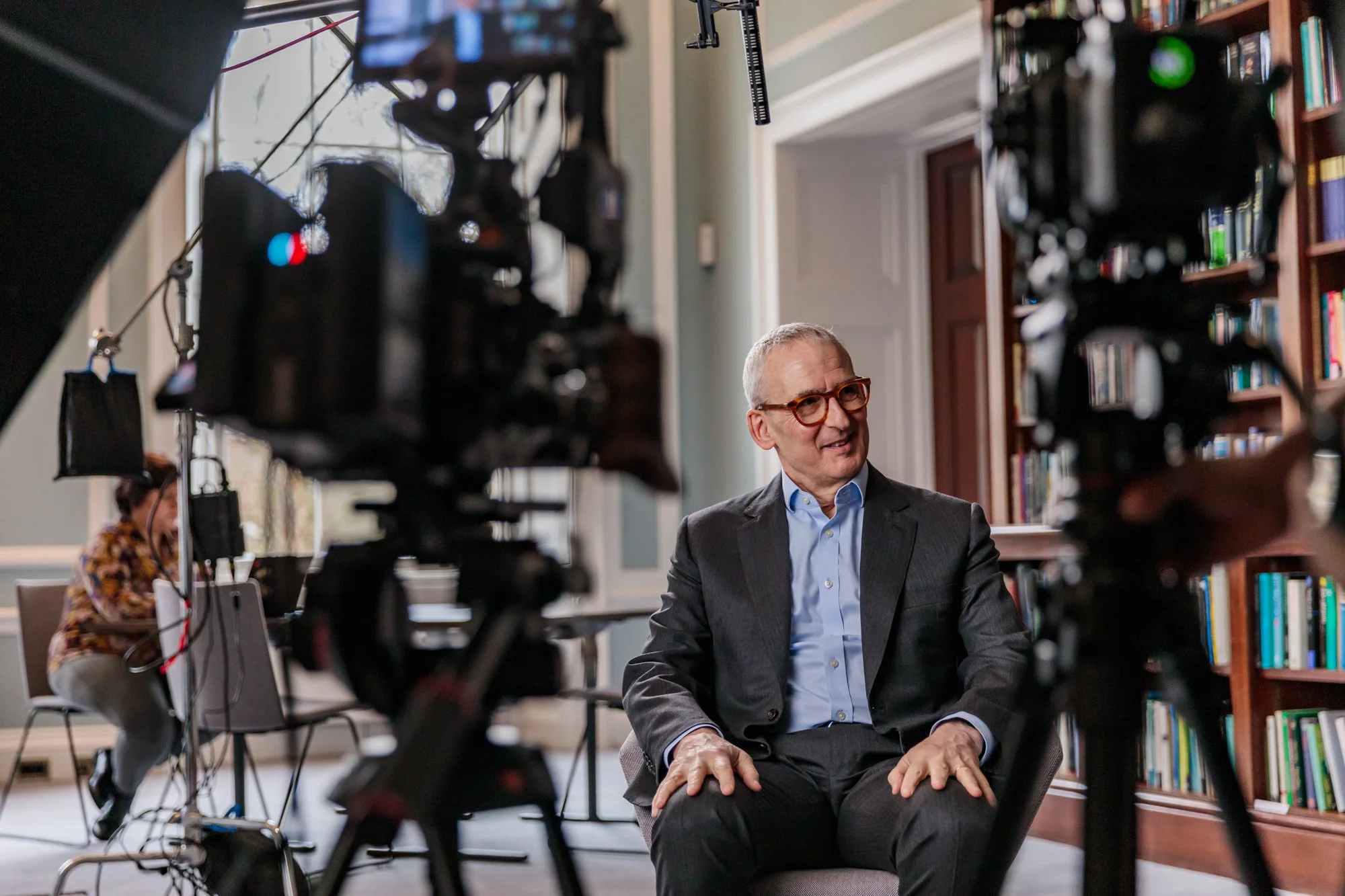 A man in a suit and glasses being interviewed or filmed in a library or office, with cameras and crew around him.